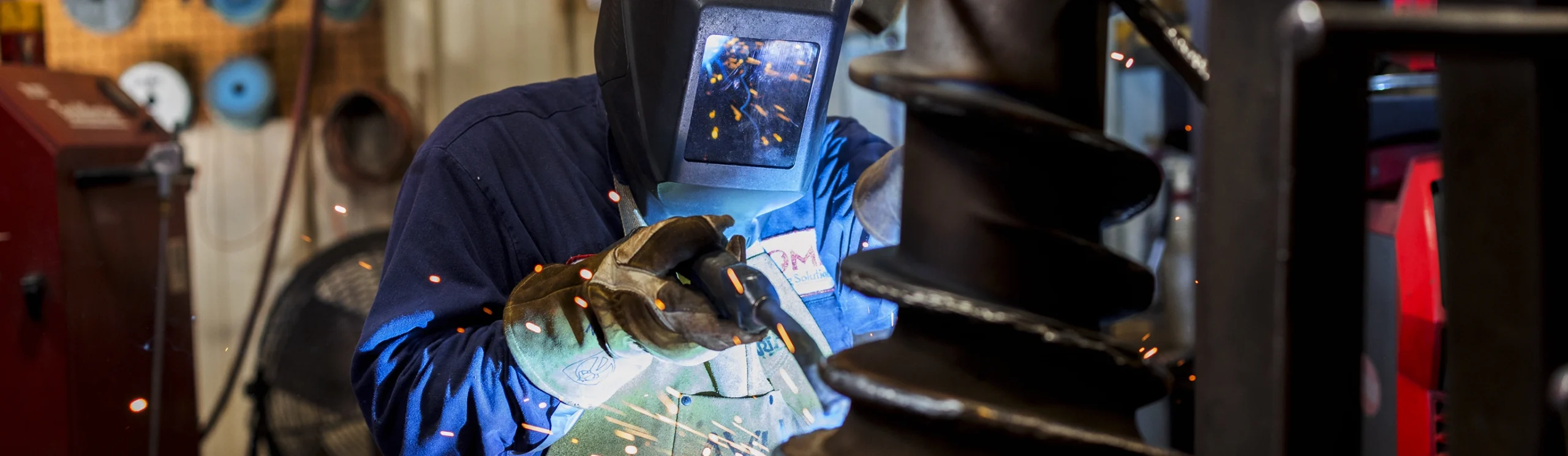 A person wearing protective gloves and a welding helmet uses a welding torch, creating sparks, inside a workshop with tools and equipment visible in the background.
