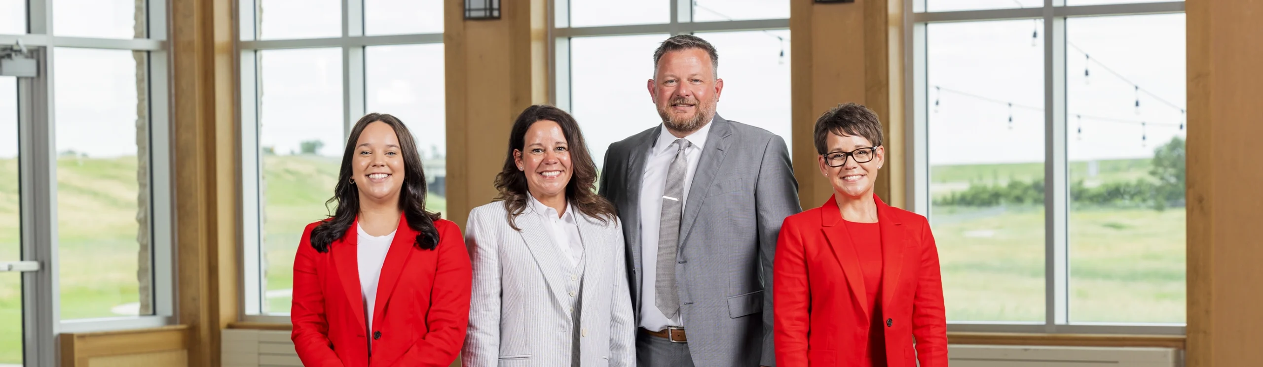 Four adults stand indoors in front of large windows; two women on the left and one woman on the right wear red blazers, and a man in a light gray suit stands between them.