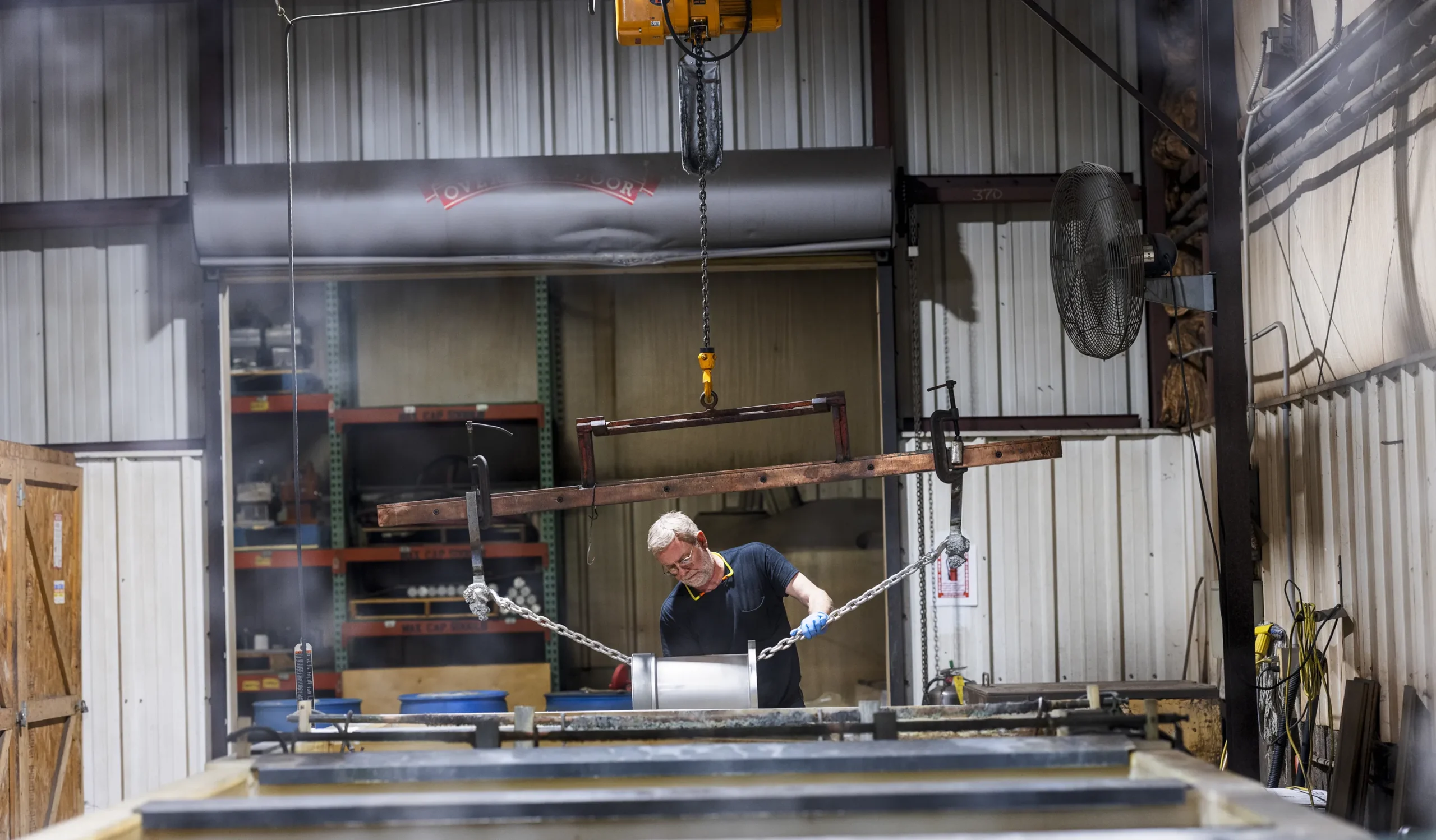 steel round shape object tighten with chains placed on working table and older man with working kit clothes working on it.